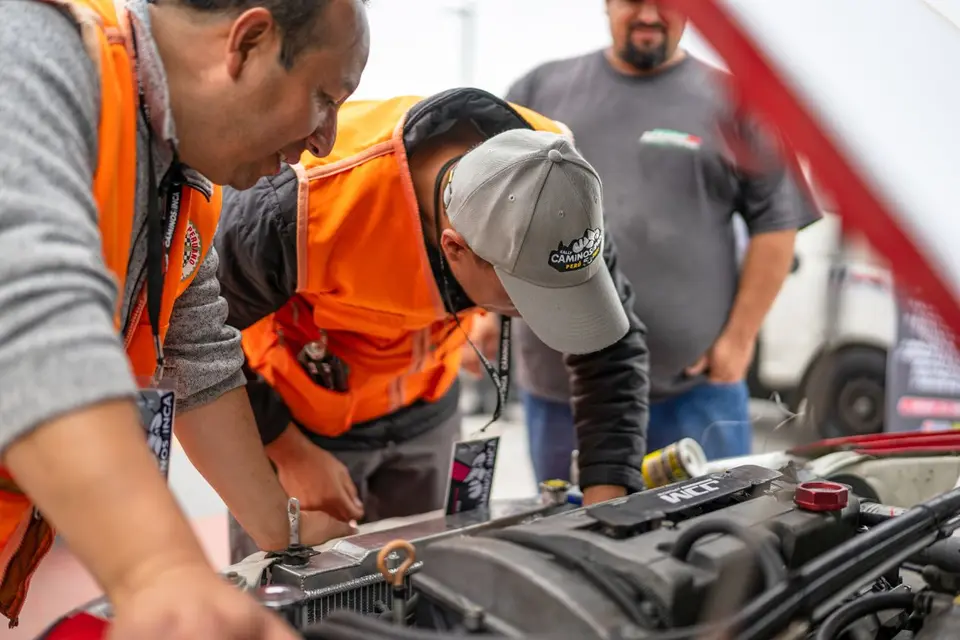 Mechanics focused on inspecting vehicle engine with open hood, wearing safety vests.
